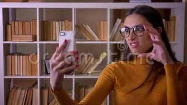 漂亮的黑发女教师<strong>戴</strong>着眼镜，在图书馆的智能<strong>手机</strong>上做滑稽和奇怪的<strong>自拍</strong>。